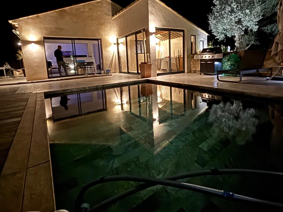 Maison avec baies coulissantes en aluminium noir, éclairée la nuit au bord d’une piscine